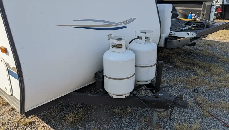 Two white propane cylinders on a travel trailer tongue with a pickup truck behind them, showing an RV propane tank repair scene.