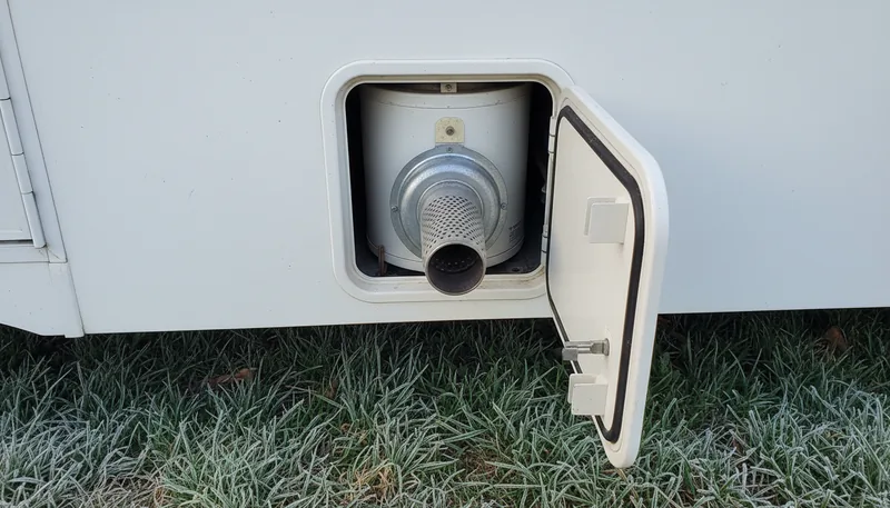 Open access door of an RV water heater with burner tube and controls on a white fifth wheel, frost on grass. RV water heater repair starts with inspecting the burner.