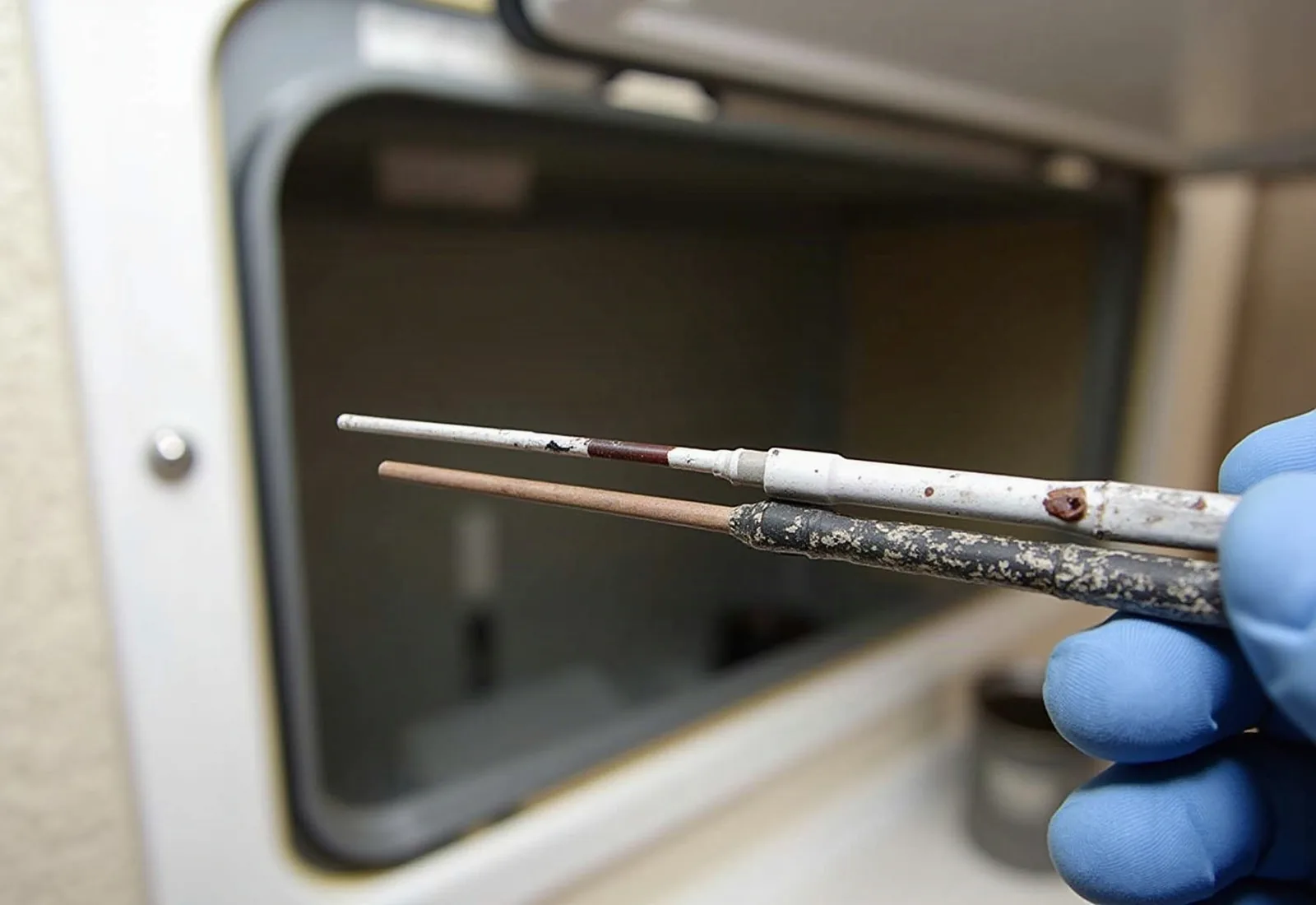 Gloved hand holding a worn magnesium anode rod next to a new one, with RV water heater access door open. Checking the anode rod is part of RV water heater repair.